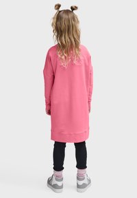 Rosa oversized sweatshirtklänning med långa ärmar, ribbade muddar och en fållad nederkant. Bärs över svarta leggings och grå sneakers med rosa detaljer.