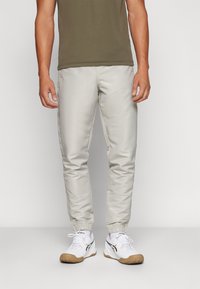 Pantalones de chándal gris claro con puños elásticos, fabricados en un tejido suave. Combinados con zapatillas deportivas blancas con suela marrón y detalles en negro.