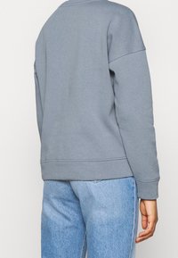 Grå sweatshirt med avslappnad passform, rund halsringning och sänkta axlar. Mjuk tyg och ribbade muddarn. Matchad med ljusblå jeans.