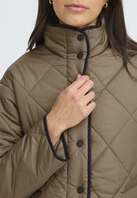 Brun kviltad jacka med hög krage, svarta detaljer och tryckknappar, som visar en slät yta och något som är sydd med diamantmönster.
