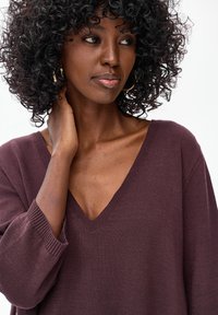 Sweter oversize z dekoltem w serek, wykonany z ciemnobordowego dzianego materiału. Posiada ściągacze na mankietach i fakturowane detale. Modelka ma kręcone włosy i złote kolczyki-koła.