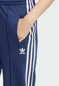 Adidas navy blue athletic pants with white side stripes, featuring a zippered pocket and a small embroidered logo on the thigh.