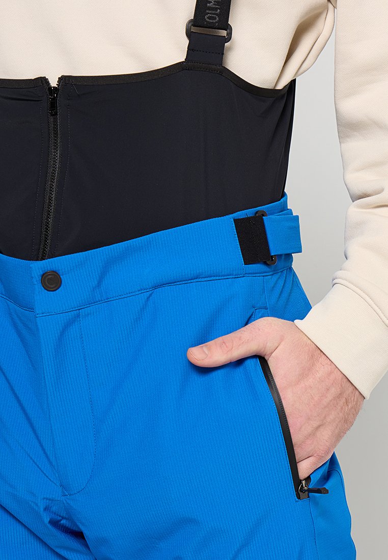 Wodoodporne niebieskie spodnie narciarskie z czarną górną częścią, regulowanymi szelkami, kieszenią zapinaną na zamek oraz wygodnym dopasowaniem w okolicy talii.