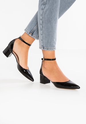 Personne portant un jean bleu clair et des chaussures à talons blocs noires en vernis avec des brides à la cheville et des bouts pointus sur un sol blanc.