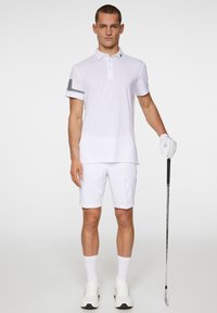 Weißes Poloshirt mit kurzen Ärmeln, verziert mit einem schwarzen Streifenakzent am linken Arm, kombiniert mit weißen Shorts und weißen Sneakern. Golfschläger wird in der rechten Hand gehalten.