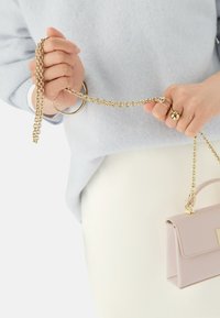 Jasnoróżowa skórzana torebka na ramię ze złotym łańcuchem, trzymana w jednej ręce. Pasek ma splecione ogniwa i dekoracyjną zapięcie.