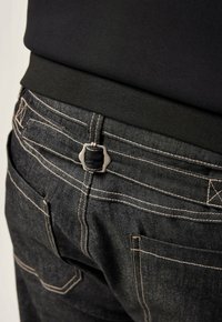I jeans di denim neri presentano un tessuto testurizzato, con cuciture chiare a contrasto e un dettaglio in metallo sul retro.