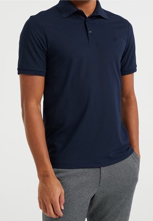 Homme portant un polo bleu marine à manches courtes et un pantalon gris à motifs, une main dans la poche, sur un fond uni.