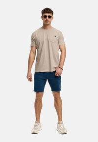 Beige T-Shirt mit kurzen Ärmeln und Brusttasche, kombiniert mit blauen Shorts und beigen Sneakers. Model trägt Sonnenbrillen und steht aufrecht.