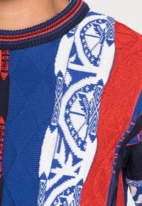 Gebreide trui met blauwe, rode en zwarte secties, voorzien van geometrische patronen, diagonale ribbeling en een gestructureerd, multicoloren ontwerp.