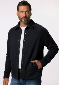 Veste boutonnée bleu marine avec un col, en tissu tricoté texturé et dotée de poches latérales. Présente des boutons-pression noirs et une coupe décontractée.