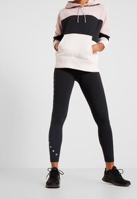 Hoodie rosa, nera e bianca, con tasca frontale. Leggings neri con scritte bianche sui lati. Scarpe sportive nere. Tessuto liscio e elastico.