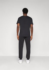 Mand med krøllet hår står med ryggen til iført en sort t-shirt, sorte joggingbukser og hvide sneakers foran en ensfarvet grå baggrund.