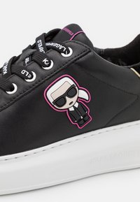 Černé kožené tenisky s robustní bílou podrážkou, černými tkaničkami a vtipným charmem v růžové a černé barvě na boku.