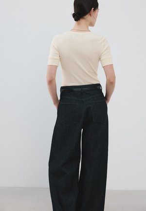 Woman with dark hair in a bun wearing a cream short-sleeve top, black wide-leg jeans, and a black belt, facing away against a plain background.