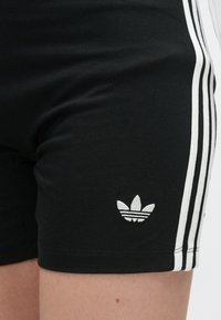 Juodi sportiniai šortai su baltomis trimis juostelėmis šonuose ir mažuoju baltu Adidas logotipu arti krašto dešinėje kojoje.