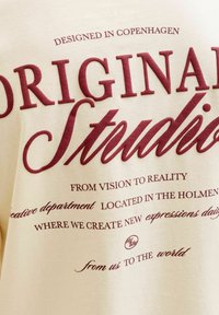 Sudadera color crema con texto burdeos. El texto incluye "DISEÑADO EN COPENHAGUE", "ESTUDIO ORIGINAL" y diversas frases en diferentes tipografías.