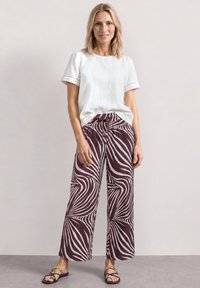 Femme debout, les mains le long du corps, portant un haut blanc à manches courtes, un pantalon large avec un motif abstrait blanc et violet foncé, et des sandales.