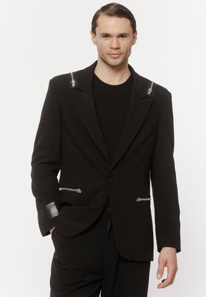 Młody mężczyzna ubrany w czarną marynarkę z suwakami na ramionach i kieszeniach, w zestawieniu z czarnymi spodniami i koszulą, na białym tle.