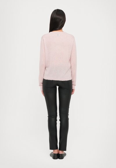 Hellrosa Langarmpullover mit leicht ausgefranstem Saum, kombiniert mit enganliegenden schwarzen Hosen und flachen schwarzen Schuhen, von hinten betrachtet.