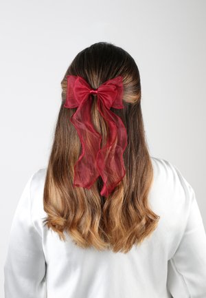 Fiocco di nastro trasparente rosso con lunghe code attaccato a capelli castani ondulati, indossato nella parte posteriore della testa, su uno sfondo bianco semplice.