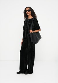 Schwarzes Samt-Zweiteiler-Outfit mit einem lockeren kurzärmeligen Oberteil und weit geschnittenen Hosen. Das Model trägt eine strukturierte schwarze Handtasche.