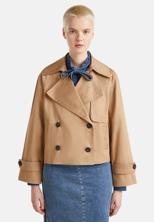 Afgebroken dubbelgeknoopte beige trenchcoat met grote zwarte knopen, brede kraag en driekwartmouwen, gelaagd over een denim rok.