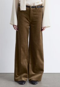 Wide-legged brown corduroy pants with a textured surface, complemented by a black belt and worn with a cream top. Black footwear visible.