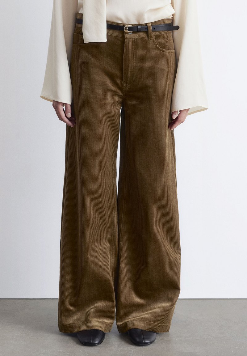 Wide-legged brown corduroy pants with a textured surface, complemented by a black belt and worn with a cream top. Black footwear visible.