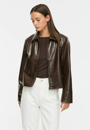 Femme portant une veste en cuir marron foncé sur un top marron et un pantalon blanc taille haute, regardant vers le bas avec de longs cheveux raides.