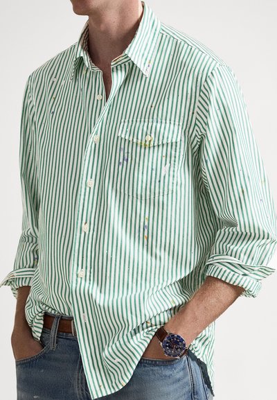 Πουκάμισο με λευκές και πράσινες ρίγες, κουμπωτό με μακριά μανίκια, με μία τσέπη στο στήθος και πιτσιλιές μπογιάς ως λεπτομέρεια.