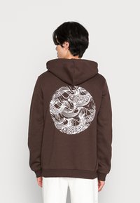 Brun hættejakke lavet af blødt stof, med et hvidt Koi fisk grafik i et cirkulært design på ryggen. Fuldt hætte med ribbede manchetter.