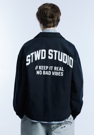 Fekete dzseki fehér "STWD STUDIO" felirattal a hátán; laza szabás és hosszú ujjú; szürke melegítőnadrágon viselve, amely szöveges részletekkel díszített.