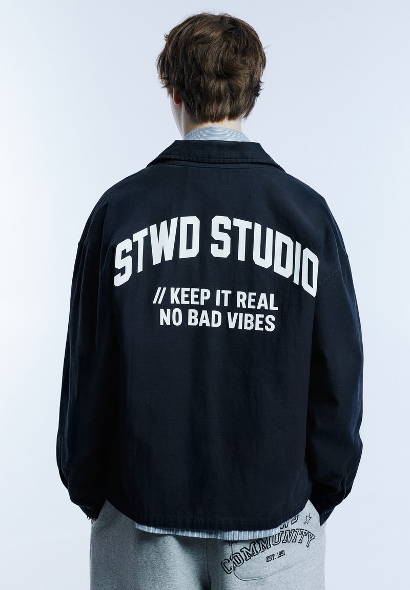 Veste noire avec le texte « STWD STUDIO » en blanc sur le dos ; coupe décontractée et manches longues ; portée sur un pantalon de survêtement gris avec un détail de texte.