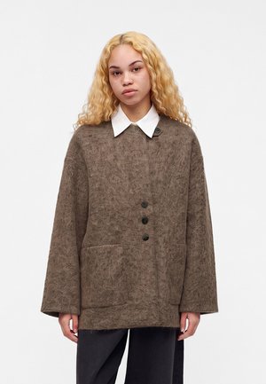 Jeune personne aux longs cheveux blonds portant un manteau marron ample texturé avec de grandes poches et des boutons noirs sur une chemise blanche à col.