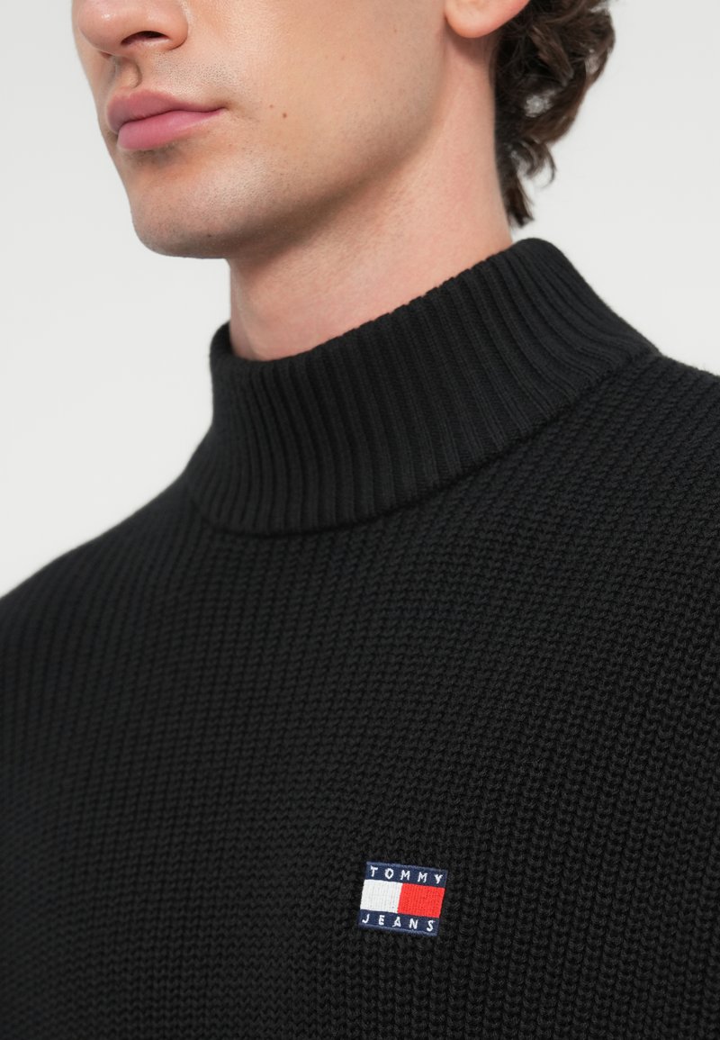 Zwarte geribbelde turtleneck trui met een nauwsluitende halslijn. Bevat een klein blauw, rood en wit "TOMMY JEANS" logo patch.
