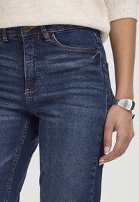Personne portant un jean en denim bleu foncé et un pull beige clair, avec un bracelet argenté au poignet droit.
