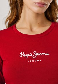 Raudonas medvilnės ilgos rankovės marškinys su baltu užrašu "Pepe Jeans" ir "LONDON" priekyje. Paprasta apvali iškirpė.