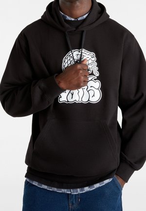 Persoon draagt een zwarte hoodie met een witte gestileerde slang en tekstlogo op de borst, hand houdt de hoodie koorden vast, blauwe spijkerbroek zichtbaar.