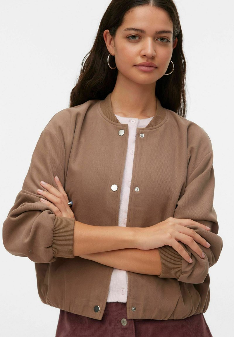 Blouson aviateur marron en tissu doux avec poignets et ourlet côtelés. Doté de boutons-pression argentés et d'un col arrondi. Superposé à un haut rose.