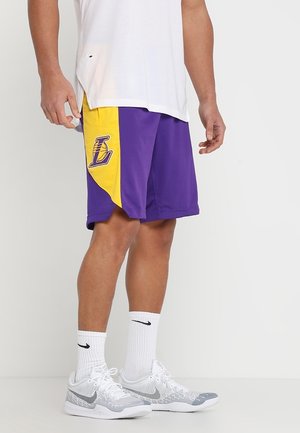 Person trägt violette und gelbe Basketballshorts mit einem "L"-Logo, weiße Nike-Socken und weiße und graue Nike-Sneaker.