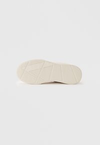 Suola di una scarpa beige che mostra modelli di battistrada geometrici con forme diagonali e triangolari su uno sfondo bianco uniforme.