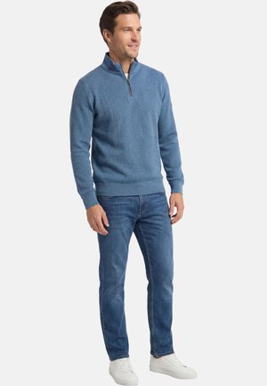 Mann trägt einen blauen Pullover mit Viertelreißverschluss, blaue Jeans und weiße Turnschuhe, steht und schaut leicht zur Seite vor weißem Hintergrund.