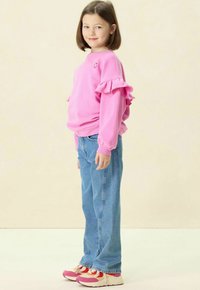 Sweatshirt rose vif avec des manches volantes, associé à un jean bleu clair et des chaussures de sport multicolores. Logo souriant simple sur la poitrine.