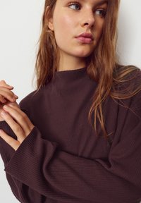 Maglia a costine marrone scuro a maniche lunghe con collo alto e vestibilità rilassata, caratterizzata da maniche lunghe con polsini allungati. Texture morbida visibile.