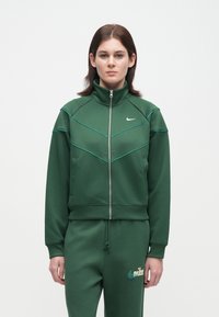 Νέος με μαλλιά μέχρι τους ώμους που φοράει μια ζακέτα με φερμουάρ σε πράσινο χρώμα της Nike και αντίστοιχες φόρμες κατά της λευκής κουρτίνας.