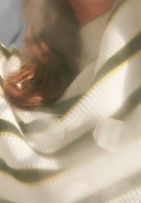 Pull blanc avec des rayures vertes et dorées, présentant une texture côtelée, un tissu drapé et une apparence douce, partiellement obscurci par un flou de mise au point.