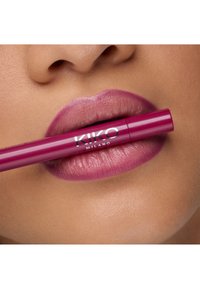 Producto labial KIKO Milano en un tubo fucsia brillante sostenido entre los labios de la modelo, con labios mauve suaves y mate que presentan líneas sutiles.