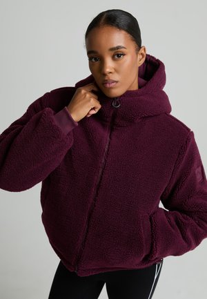 Mujer con una chaqueta de forro polar burdeos con cremallera y capucha, mano cerca del cuello, sobre un fondo gris liso.