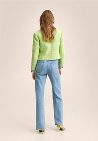 Femme avec de longs cheveux ondulés portant une veste texturée vert clair, un jean droit bleu clair et des talons ouverts jaunes, se tenant sur un fond neutre.